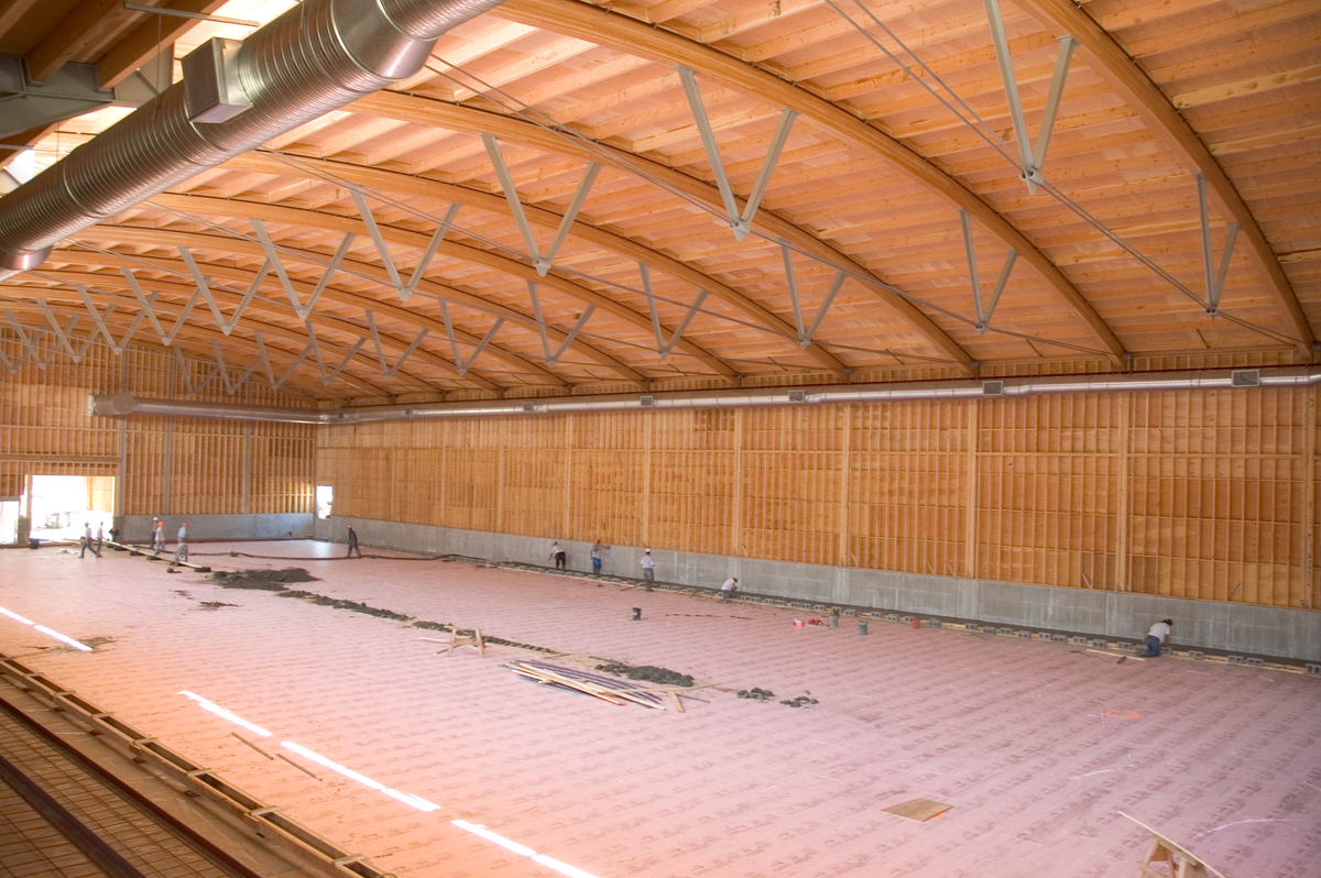 Armstrong-Spallumcheen竞技场的室内白天视图显示了拱形胶合层合(胶合)梁，上面是木桁架和胶合板屋顶，下面是预制木面板墙