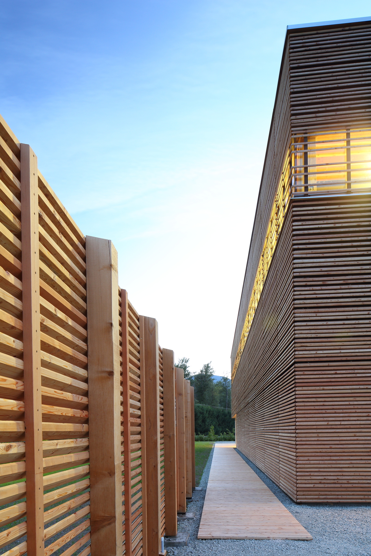 BC被动式房屋工厂的下午外部景观，这是一座低矮的被动式房屋结构，由轻质框架和大量木材构件建造，展示了围绕外部木条、木制走道和装饰性木栅栏的包裹