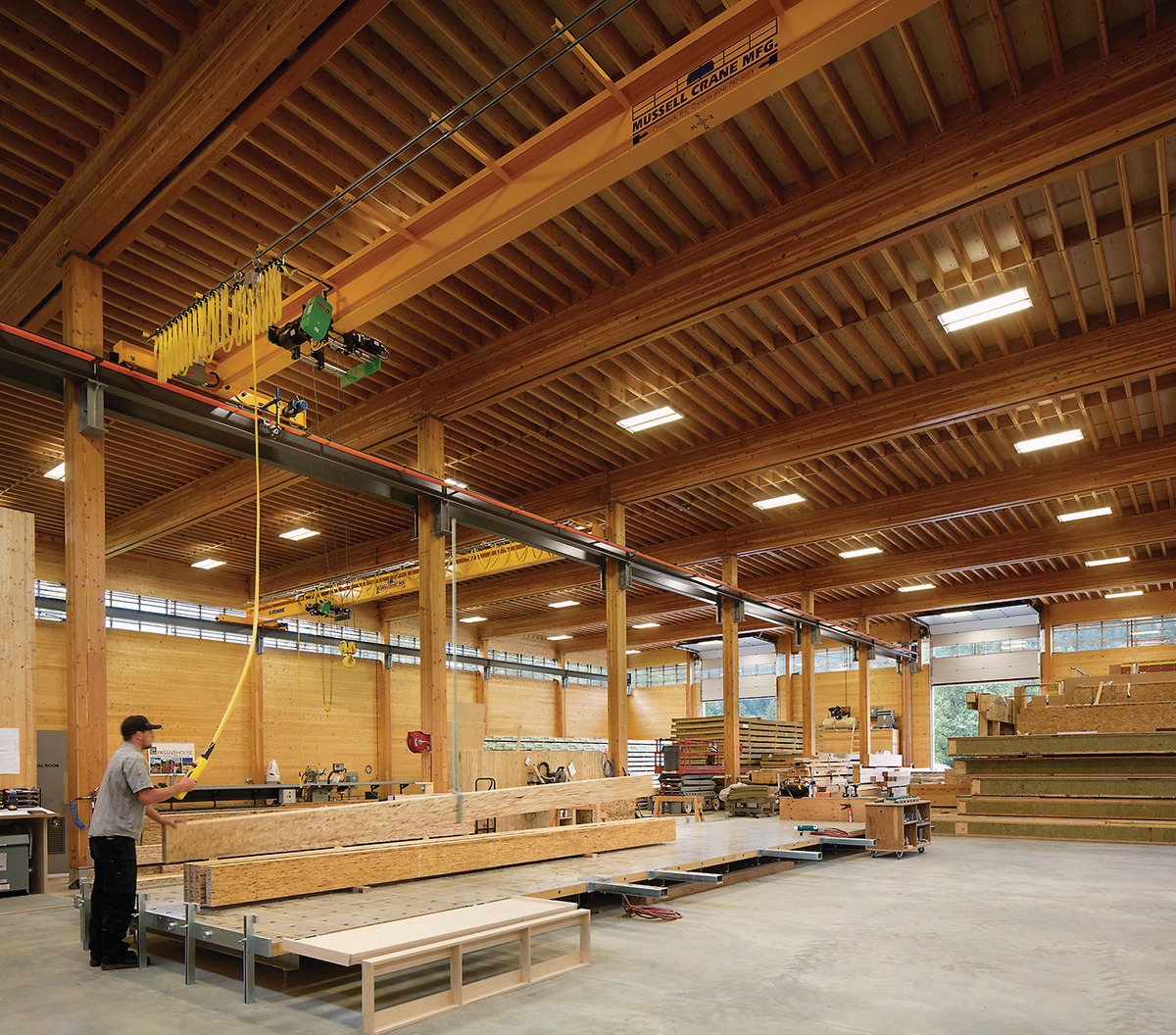 BC被动式房屋工厂的内部视图，显示工人使用桥式起重机移动大量木材梁