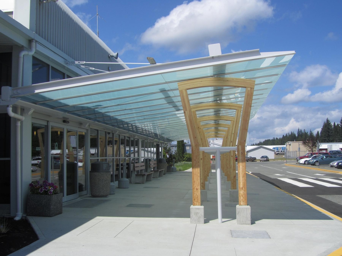 坎贝尔河机场扩建项目低层有盖入口的室外阳光白天视图，显示胶合木“拱门”结构，支撑着一个大型玻璃雨棚，创造了一个室外入口房间