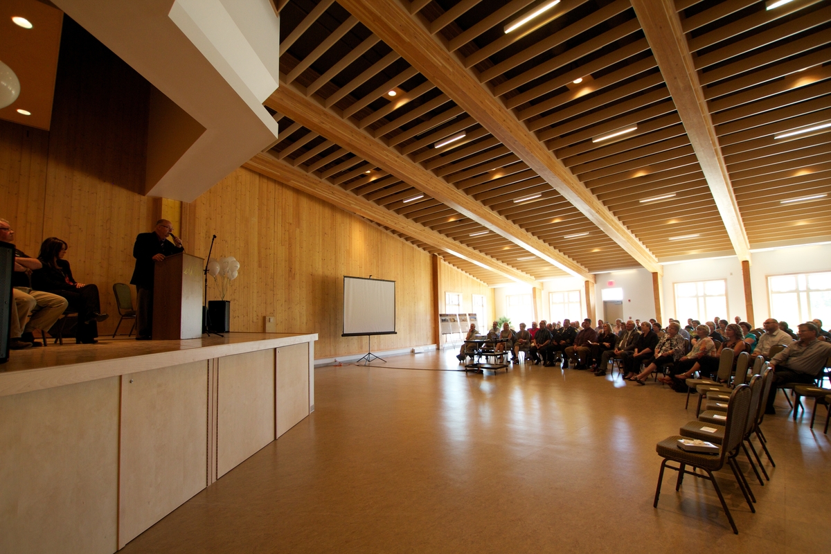 在Elkford社区会议中心主厅内，坐在讲台上的与会者和演讲者的室内日间视图，大厅包括胶合层压木材(Glulam)和层压单板木材(LVL)梁的结构元素，以及交叉层压木材(CLT)，提供可见的墙壁和天花板/屋顶结构