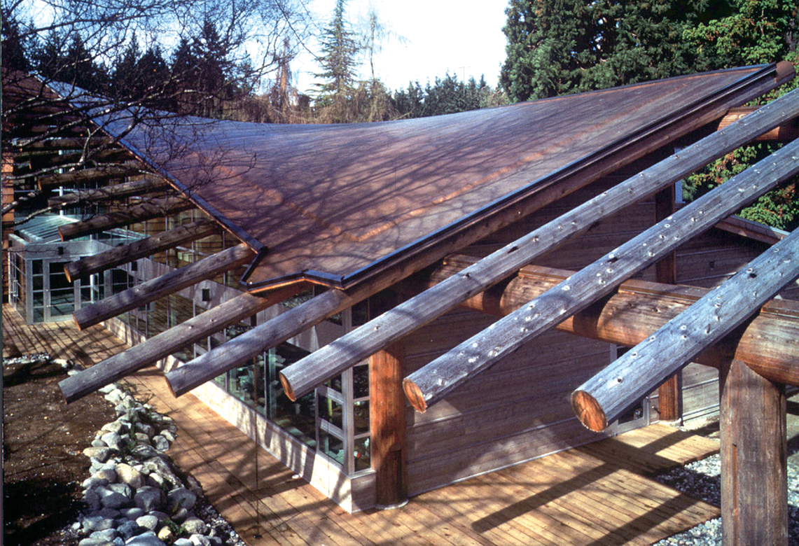 英属哥伦比亚大学第一民族长屋的户外日间航拍图像，显示玻璃和木材外部，顶部是大量木材实心锯重木材屋顶结构，设计成类似飞行中的鸟的翅膀