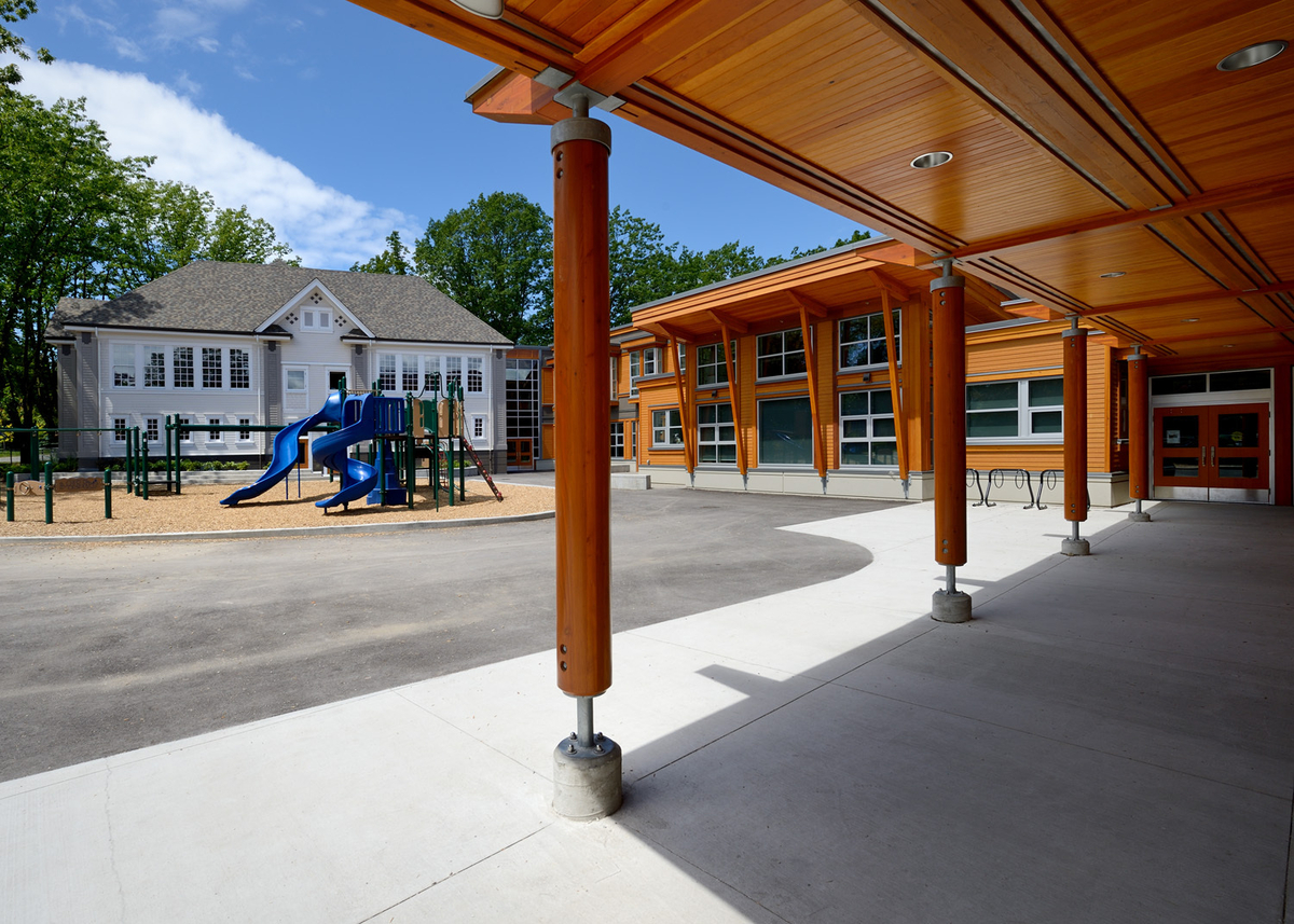 基奇纳勋爵小学白天阳光明媚的外部图像拍摄于主入口的木制外部屋顶下，显示了木材的广泛使用，包括胶合木(胶合木)、木制品、镶板和壁板