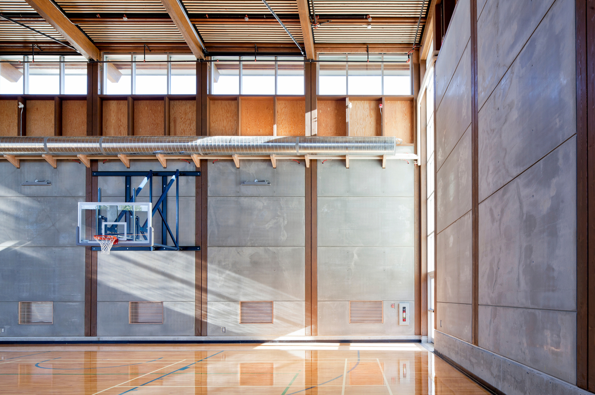 奥卡那根学院吉姆·帕蒂森卓越中心体育馆低层的室内白天视图，展示了混合木材和混凝土结构，包括胶合层压木材(胶合木)屋顶梁