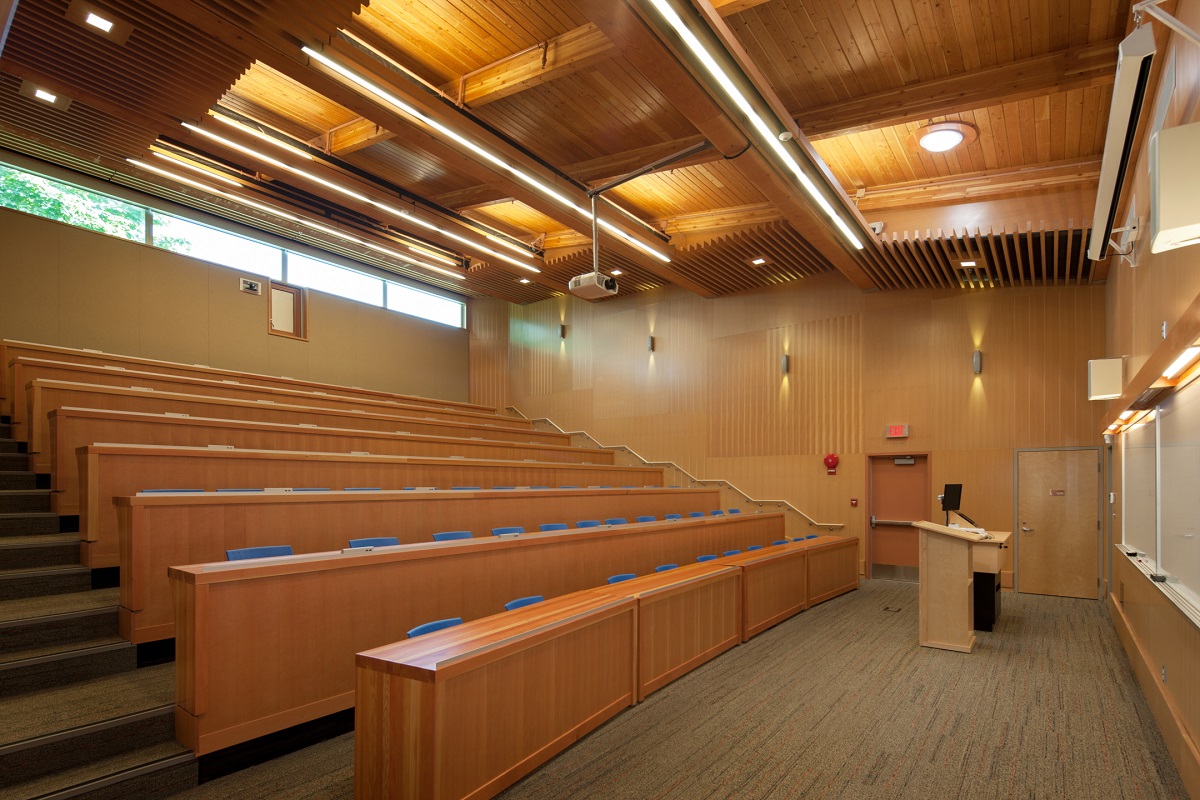 胶合木(胶合木)梁支撑舌和槽天花板木材都展示在温哥华岛大学考伊肯校区报告厅的白天室内图像中，配有蓝色塑料椅子的木制柜台配置为礼堂座位