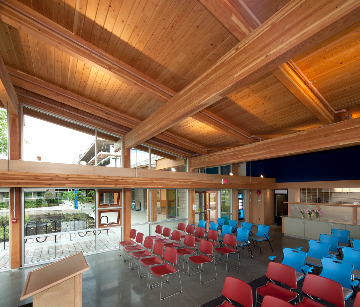 胶合木(胶合木)梁支撑舌和槽屋顶木材都展示在温哥华岛大学考伊肯校区多层主礼堂的白天室内图像中，配有红色和蓝色的塑料椅子