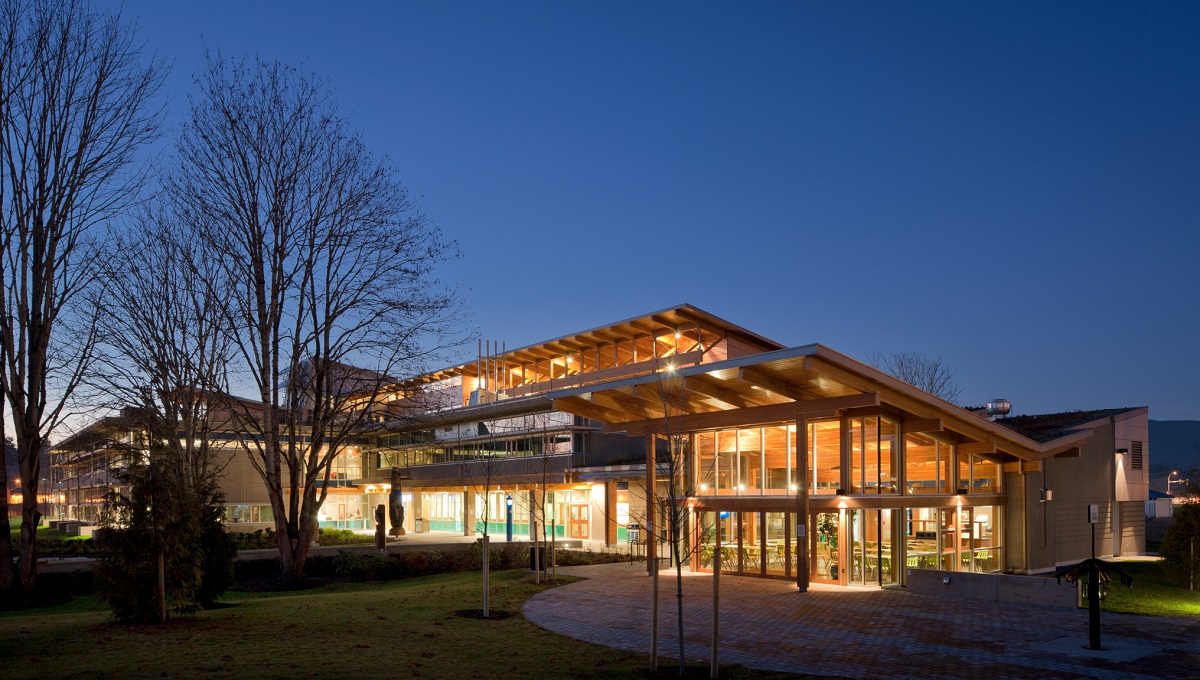温哥华岛大学考伊肯校区的外部建筑夜景，展示了明亮的玻璃建筑前面，包括胶合木(胶合木)梁和柱，支撑着膨胀的木质屋顶