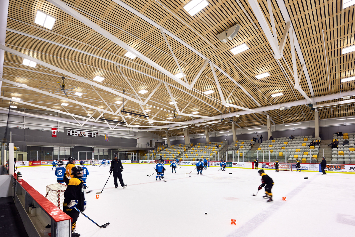 西弗雷泽中心溜冰场的内景展示了冰球训练，突出了主场馆的大型屋顶结构，框架是弯曲的钢梁桁架，由木板条天花板组件强调