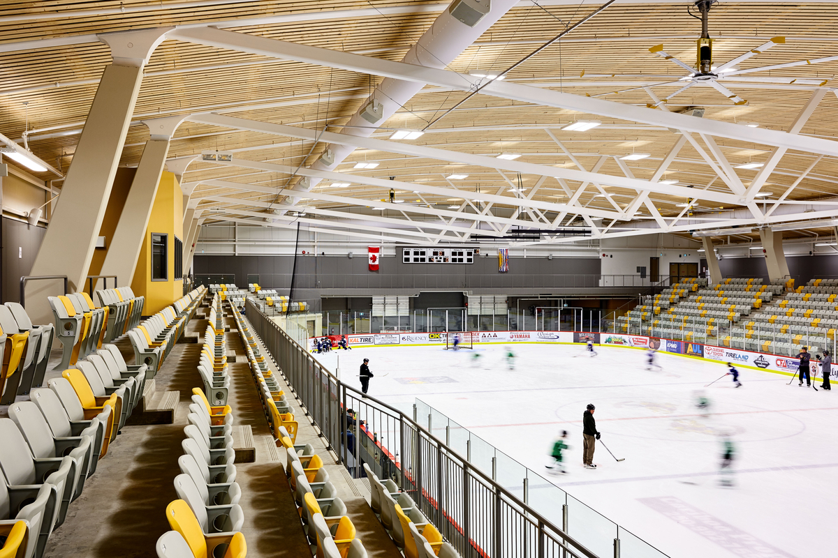西弗雷泽中心冰场的室内上层座位视图展示了曲棍球练习，并突出了主场馆的大型屋顶结构，由弯曲的钢梁桁架组成，由木条天花板组件强调