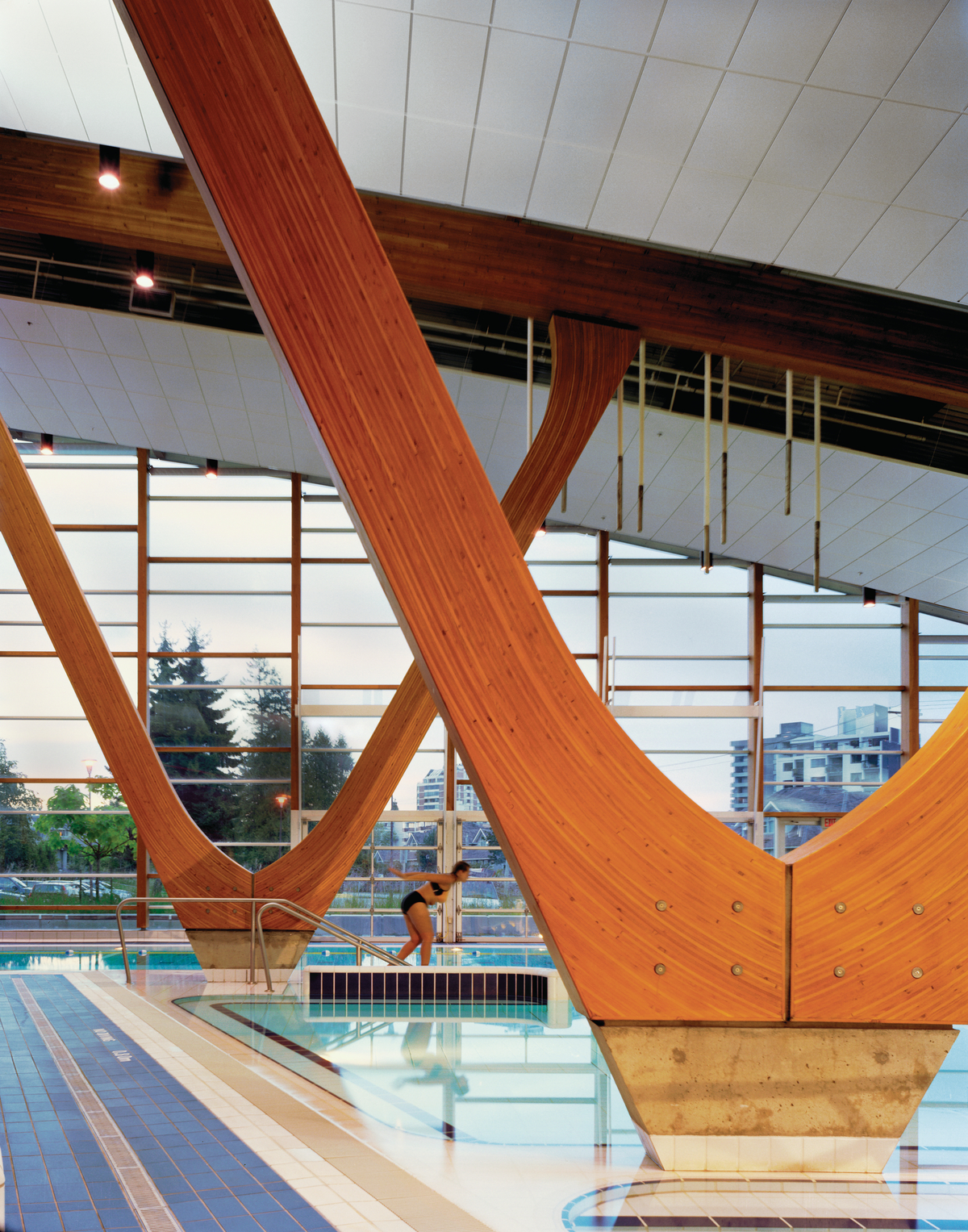 西温哥华水上中心低层建筑的室内日间景观，显示泳池表面、游泳者和弯曲的胶合木(胶合木)柱，其弧线向上支撑胶合木(胶合木)屋顶梁