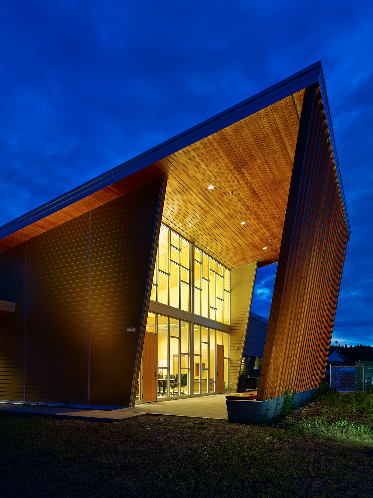 YunesitIn健康中心入口的外部夜景显示了广泛的木材和玻璃的使用，包括木板壁板和胶合木(胶合木)柱支撑预制木材屋顶桁架