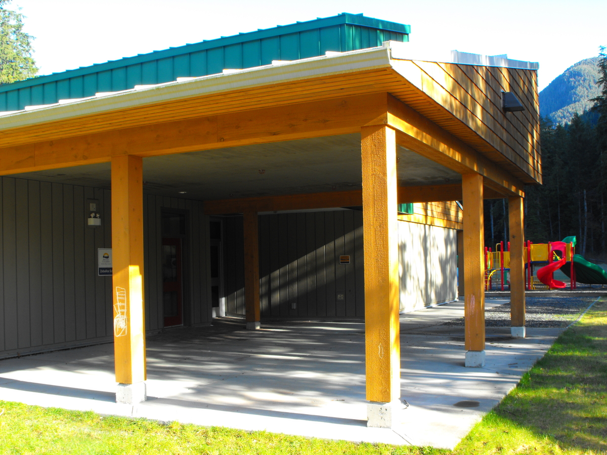 Zeballos小学/中学的白天外部图像，显示木质覆盖的入口区域，实心锯重木柱，木材天花板和木质屋顶瓦