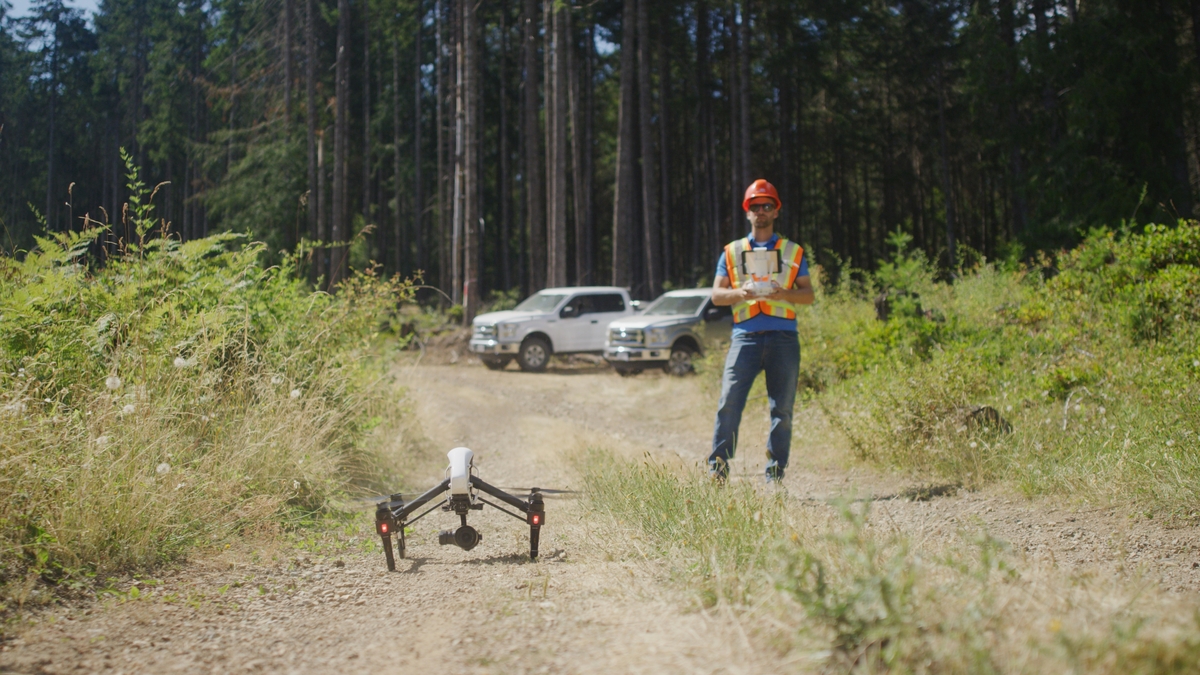 户外阳光图像林业调查工人在PPE站在土路与森林区域的背景，无人机在起飞位置，和控制器在手中